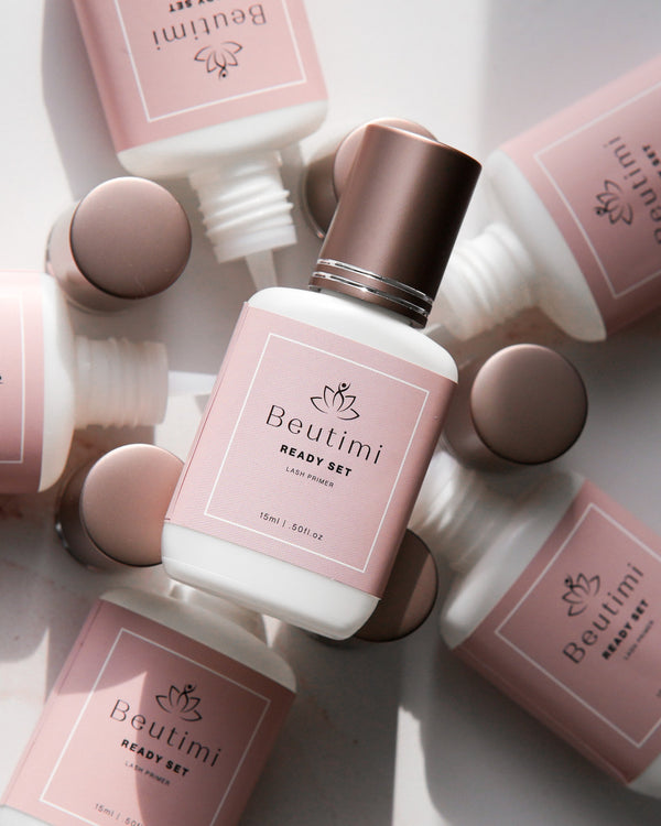 Beutimi Ready Set Lash Primer bottle with bronze cap surrounded by pink packaging and applicator tools on white surface