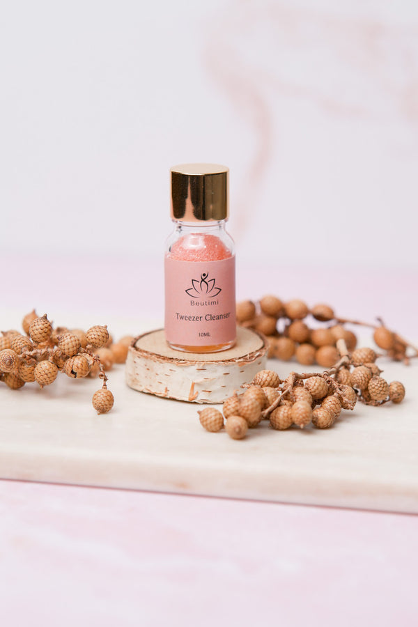 Beutimi Tweezer Cleanser in a pink 10ML bottle with gold cap displayed on birch wood surrounded by dried botanicals