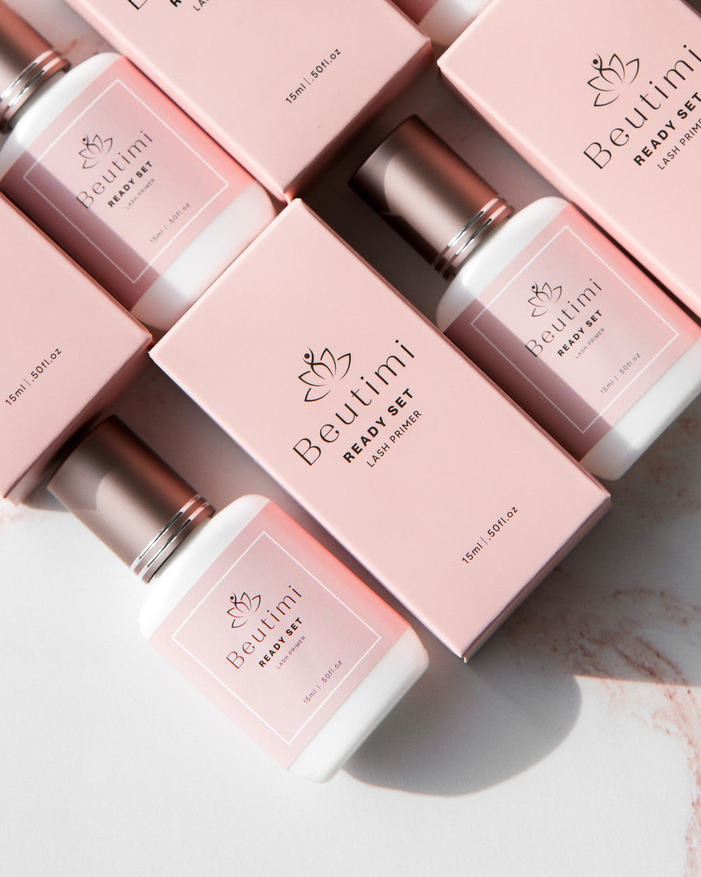 Beutimi Ready Set Lash Primer bottles in pink packaging arranged on white surface with shadows