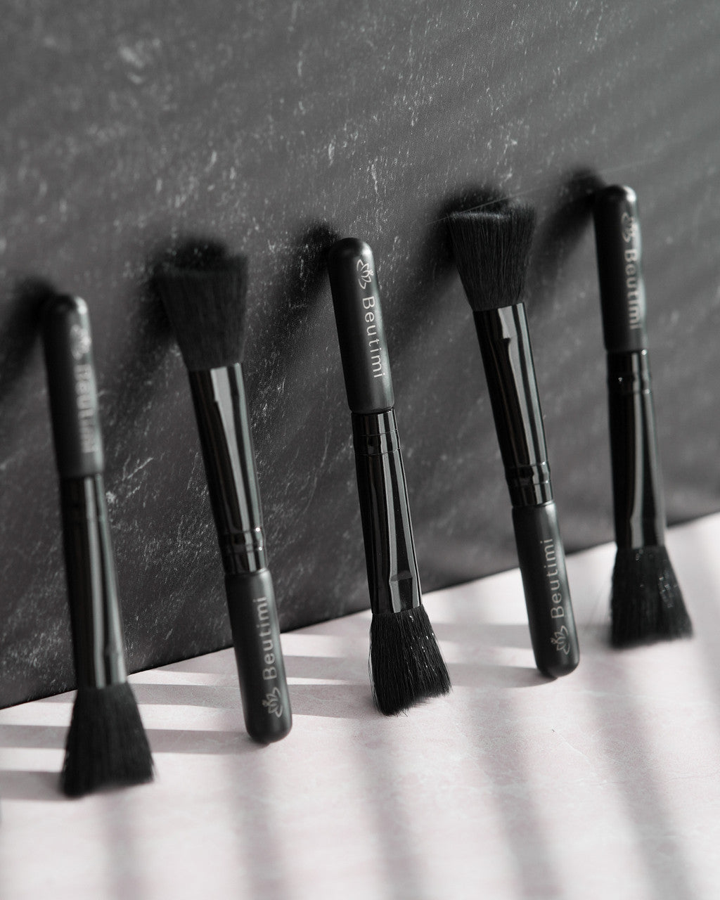 Five black Beutimi Cleansing Brushes with soft bristles arranged in a row against a textured gray backdrop in monochrome