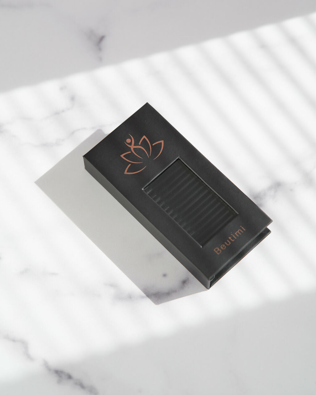 Beutimi FauxMink Lashes 0.05MM black packaging with copper lotus logo displayed on marble surface with window shadows