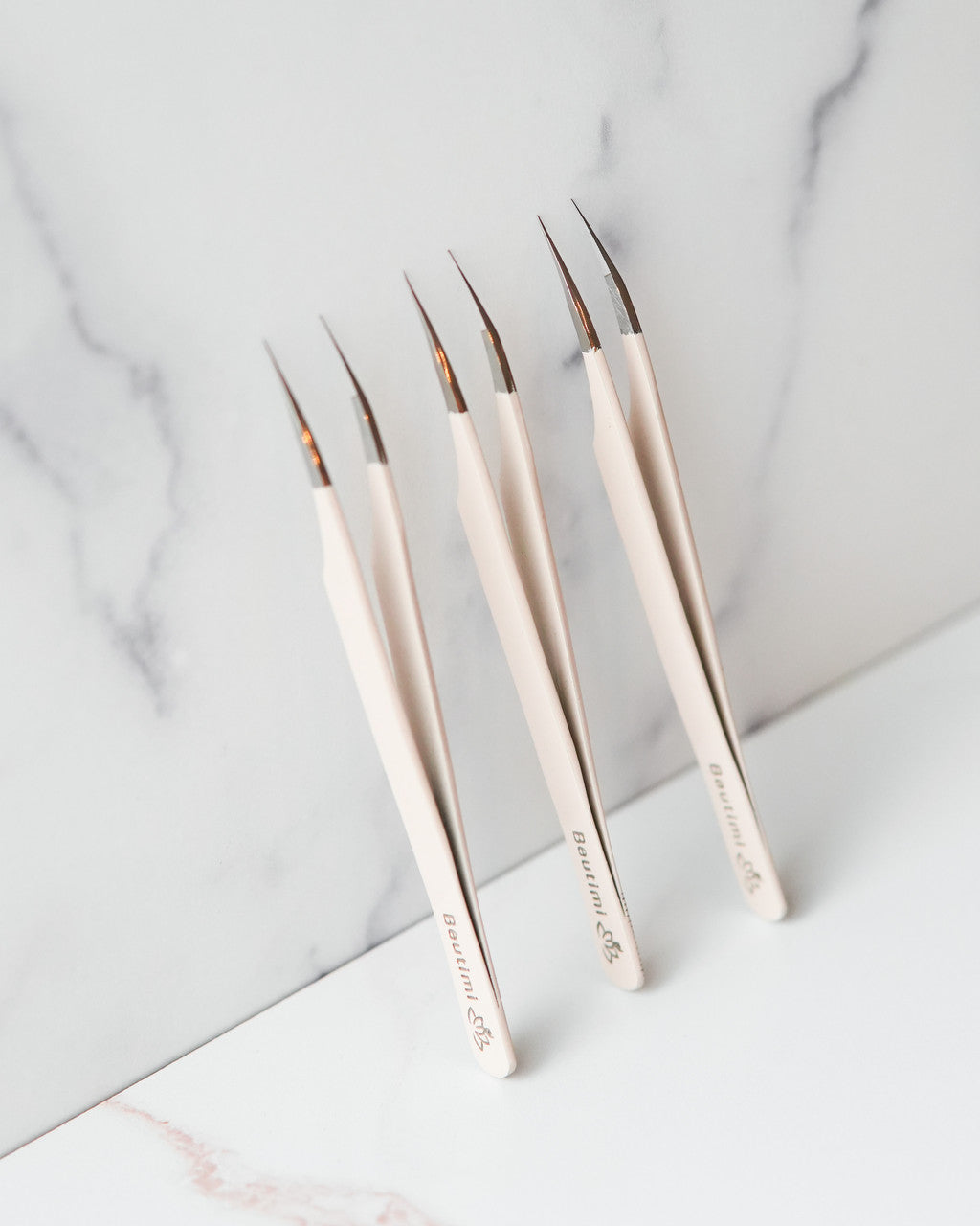Beutimi Thrive Tweezer set of six displayed at angle showing cream handles with fine dark tips on marble background