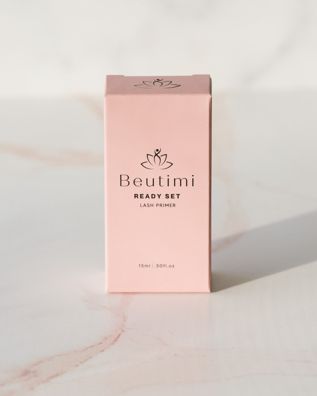 Beutimi Ready Set Lash Primer in a pale pink rectangular box with lotus logo displayed against white marble surface
