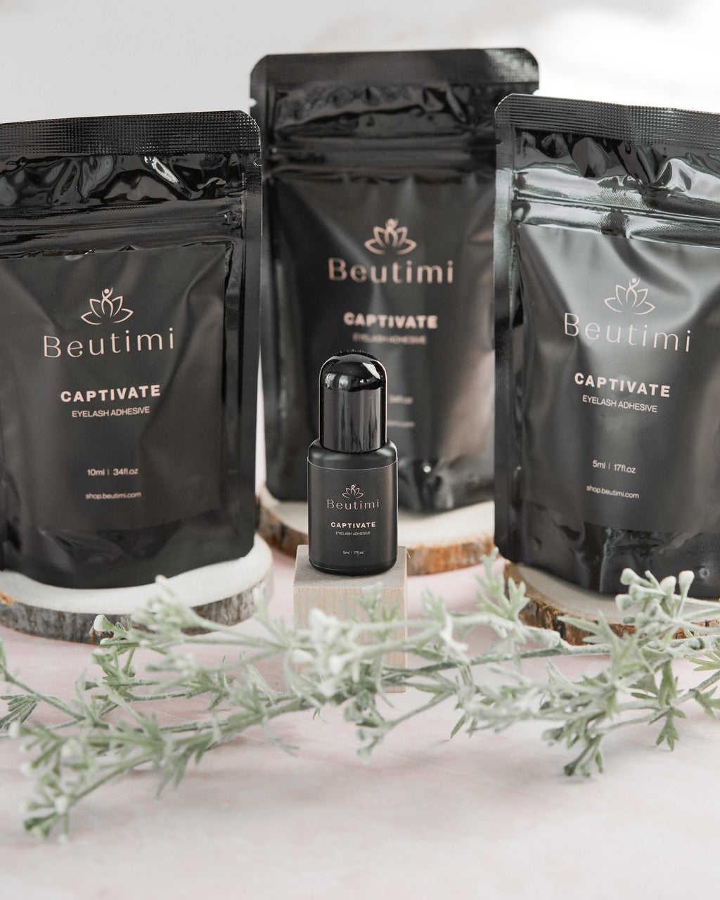 Beutimi Captivate Adhesive bottle displayed with black pouches and greenery on pink and wood surface