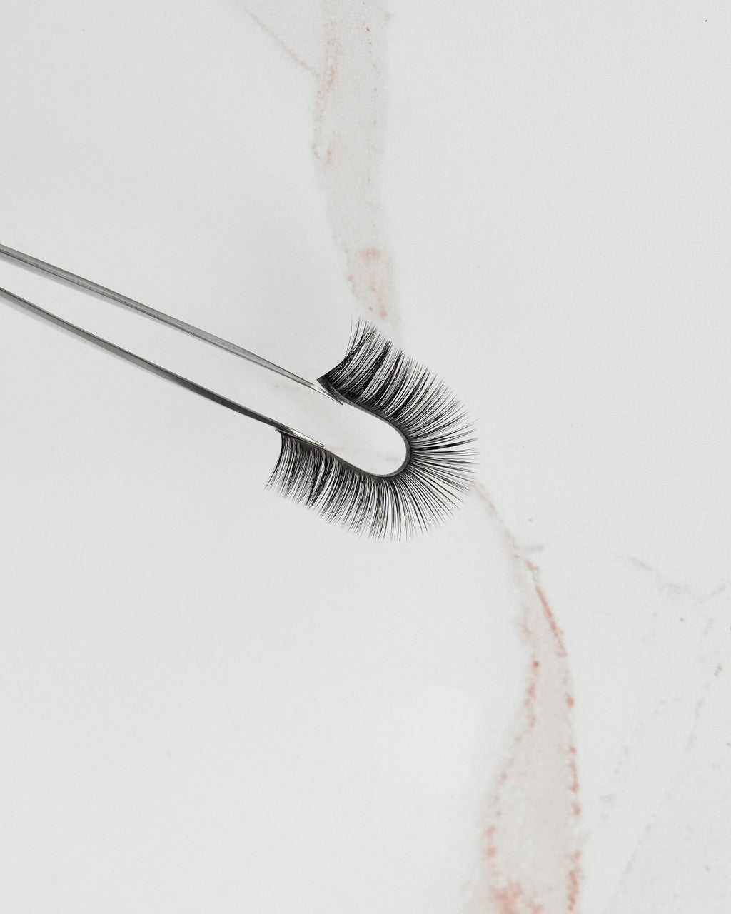 Beutimi FauxMink Lashes 0.02MM held by tweezers against marble, showcasing voluminous black lashes with deep matte finish