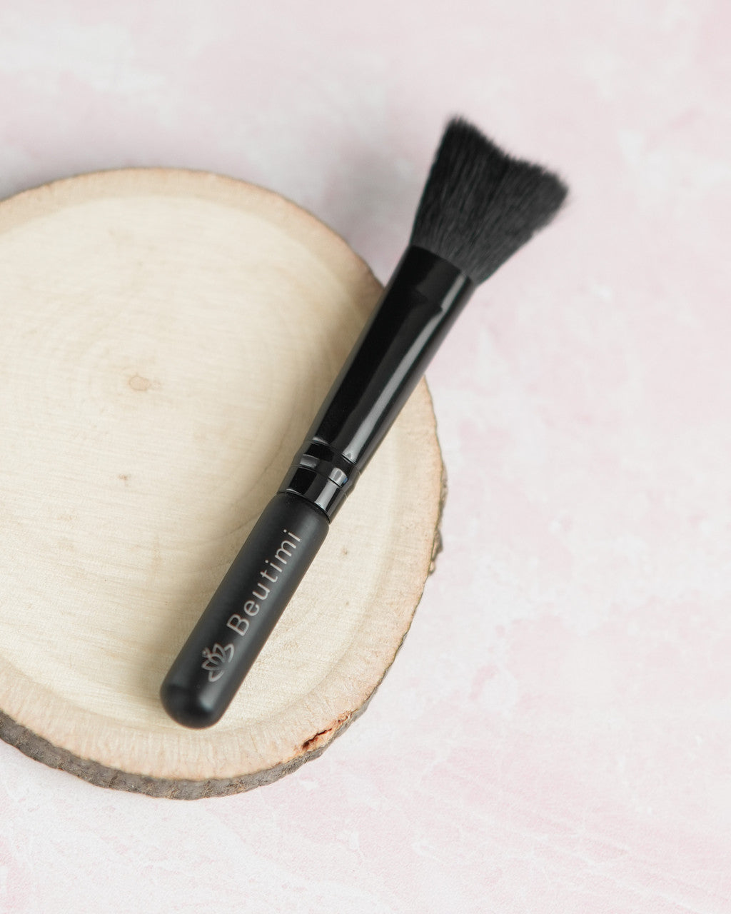 Beutimi Cleansing Brush with soft black bristles displayed on a wooden disc against a pink background
