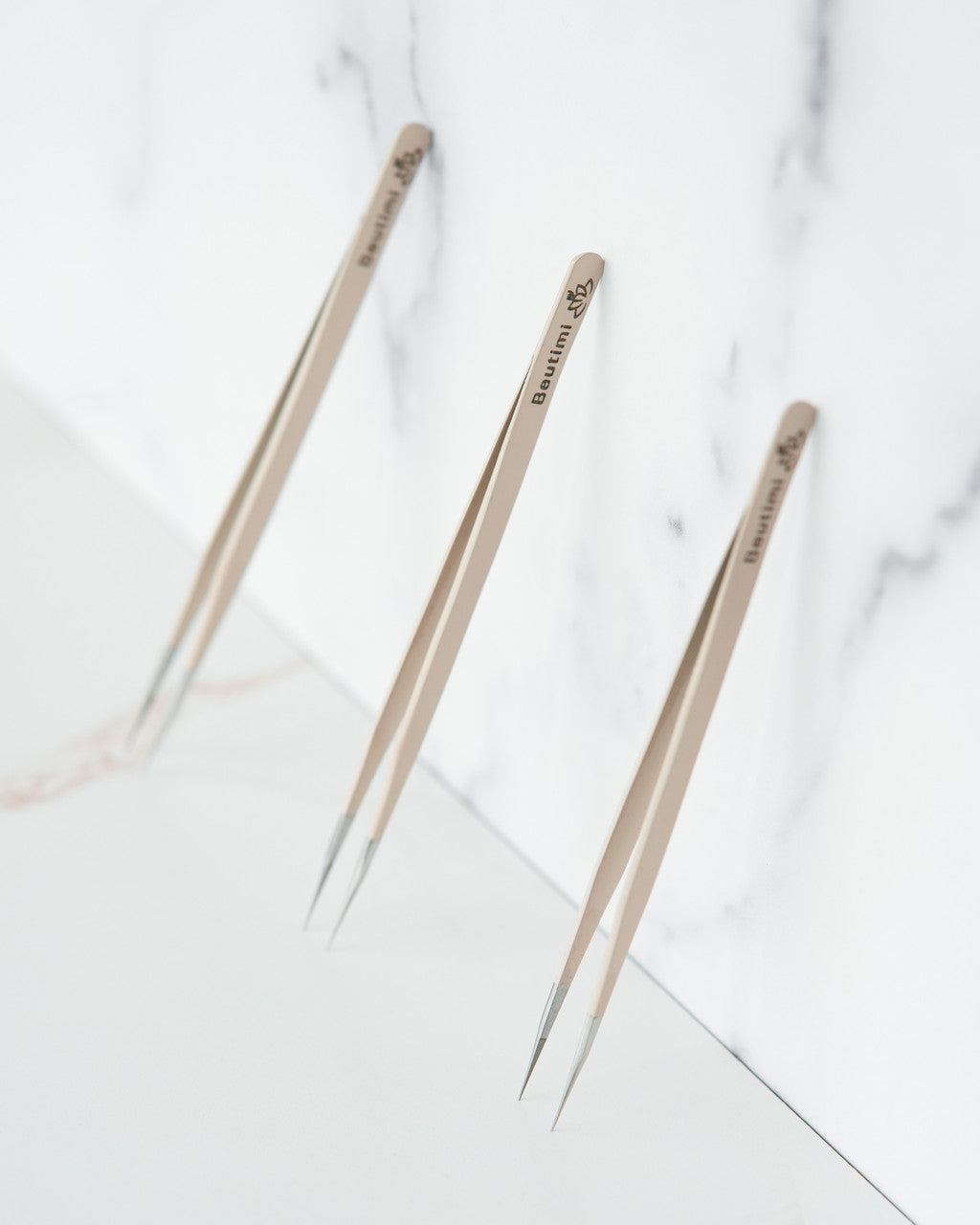 Three Beutimi On Point Tweezers with beige handles and fine metal tips arranged on a marble surface