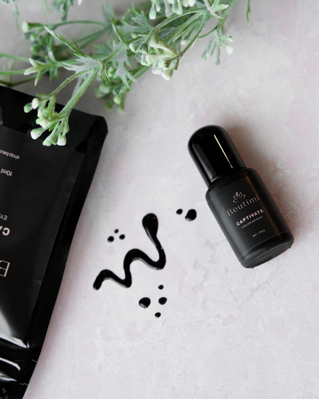 Beutimi Captivate Adhesive dark bottle displayed with swatches on white surface beside greenery and black packaging