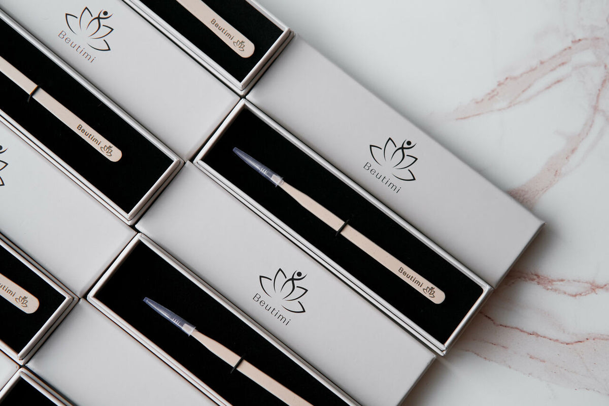 Beutimi On Point Tweezers displayed in white and black packaging arranged diagonally on pink marble surface