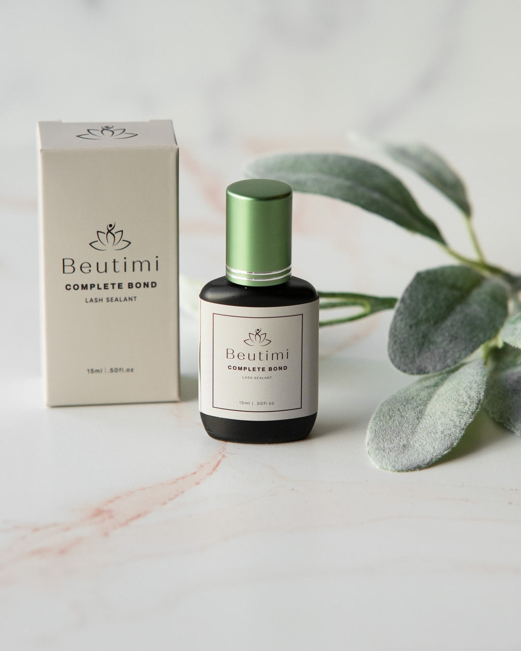 Beutimi Complete Bond lash sealant bottle with green cap displayed next to cream packaging and eucalyptus leaves on white surface