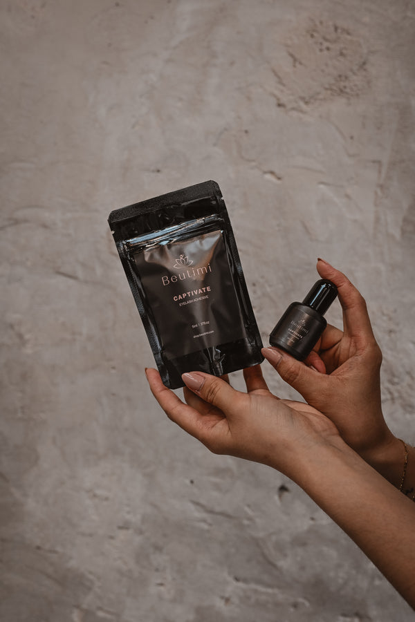 Hands holding a black Beutimi Captivate Adhesive pouch and small bottle against a textured white background