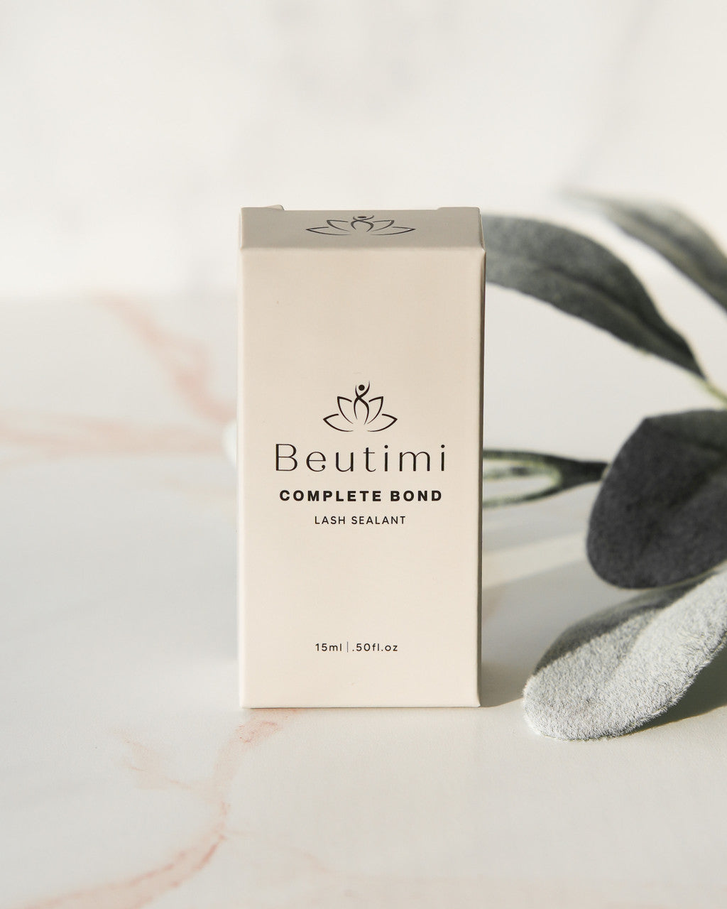 Beutimi Complete Bond lash sealant in cream packaging with lotus logo, displayed next to green plant leaves and white applicator brush