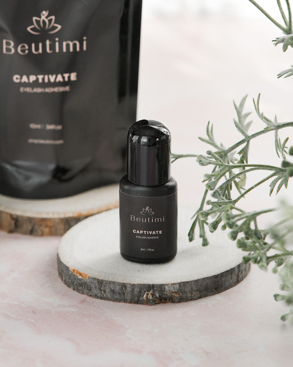 Beutimi Captivate Adhesive in a black 5ml bottle displayed on a wooden slice with green foliage
