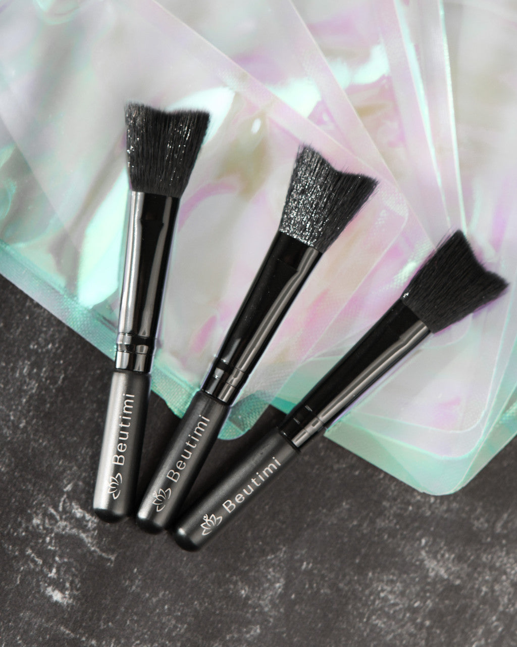 Three black Beutimi Cleansing Brushes with soft bristles displayed on a textured surface against pastel fabric background