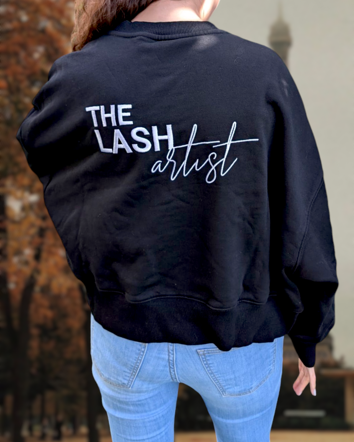 Beutimi Lash Artist Sweater in black with white text worn with light blue jeans against a brick wall