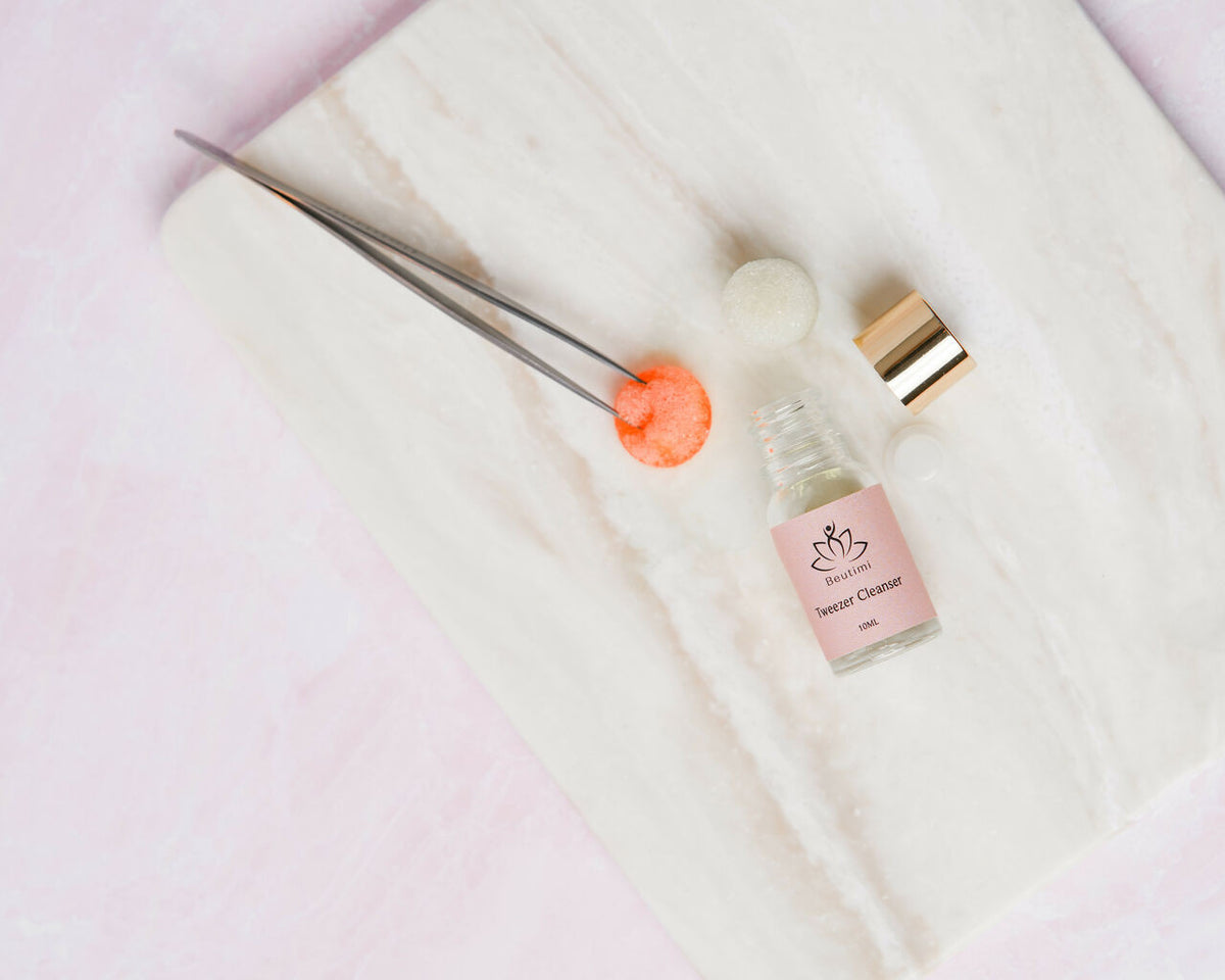 Beutimi Tweezer Cleanser bottle with pink label displayed on white cloth alongside metal tweezers and cleaning tools