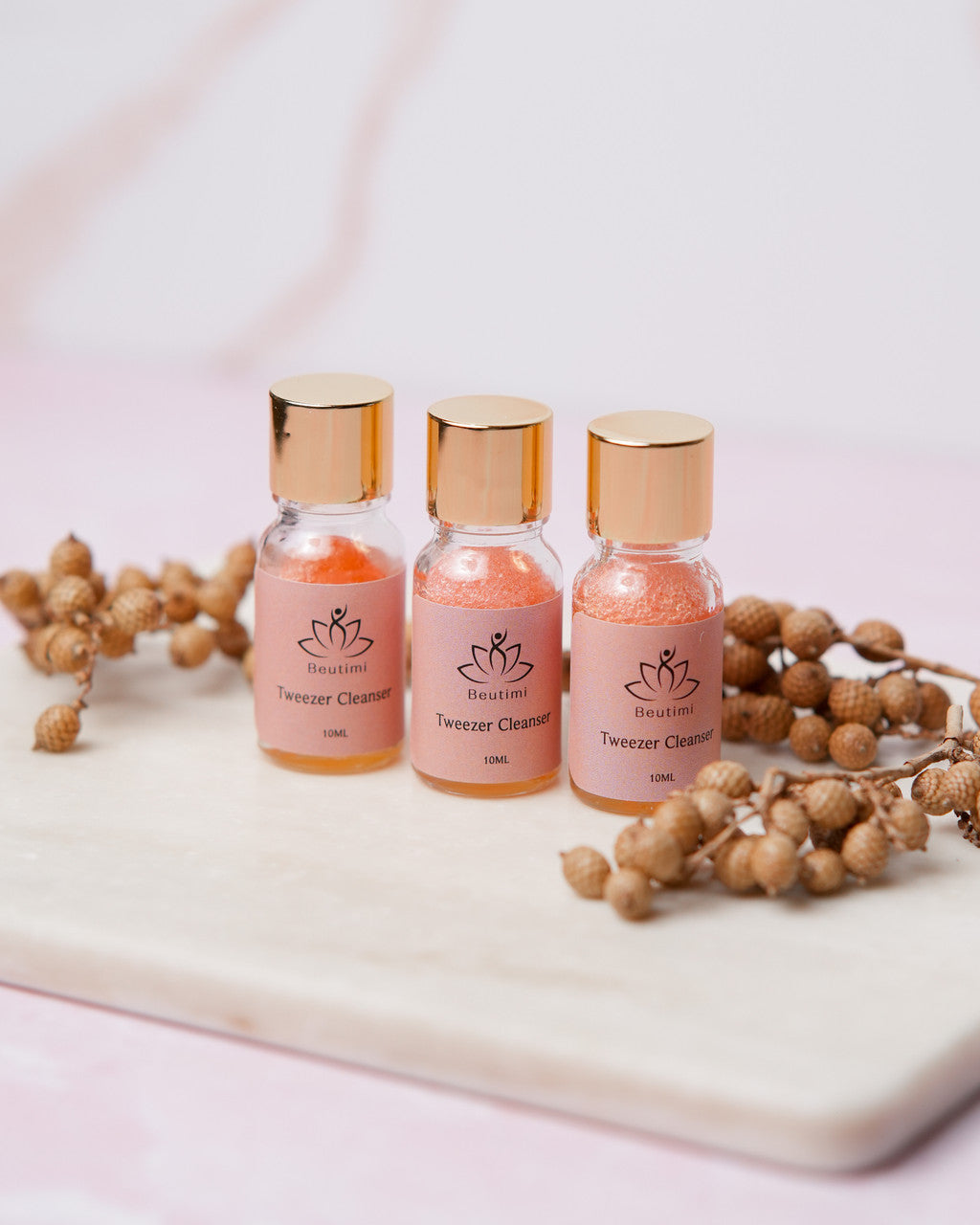 Three Beutimi Tweezer Cleanser bottles with rose-gold caps displayed on white marble with dried botanicals