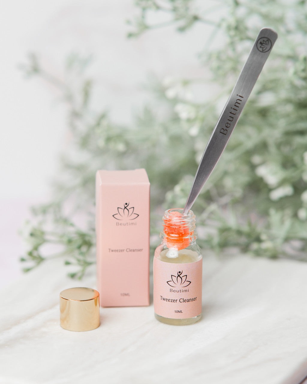 Beutimi Tweezer Cleanser in pink bottle with brush applicator, gold cap, and packaging displayed against floral background