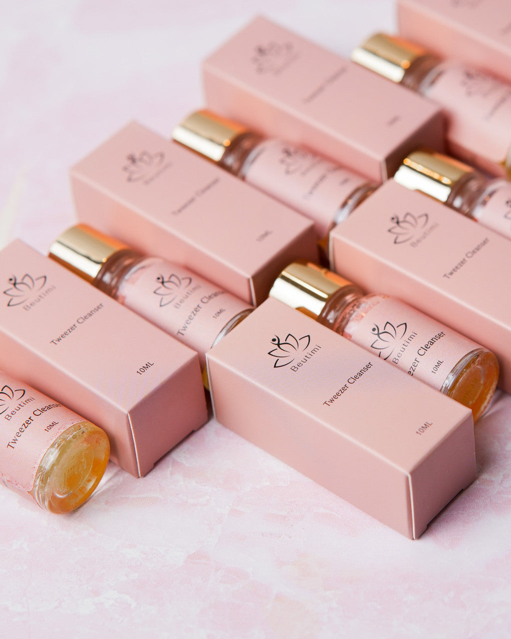 Multiple pink boxes of Beutimi Tweezer Cleanser with amber glass bottles and gold caps arranged on a light surface